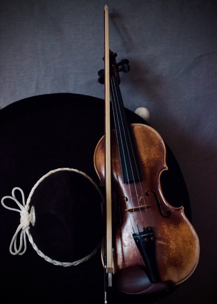 Primer plano artístico de un violín de madera clásica junto a un sombrero de mariachi negro sobre fondo oscuro.