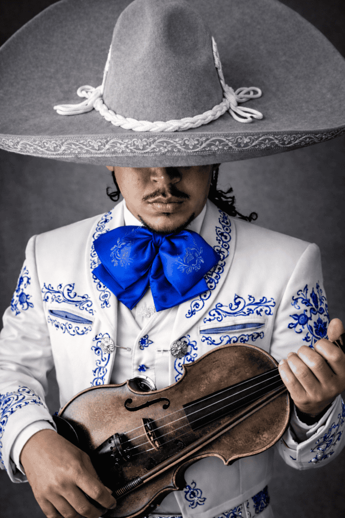 Retrato artístico de un mariachi con traje de charro blanco y bordados azules, sosteniendo un violín en una composición de color selectivo.