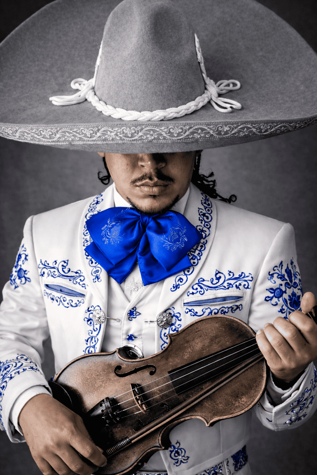 Mariachis en Alicante | Serenatas, Bodas y Eventos 24 Horas 76 Retrato artístico de un mariachi con traje de charro blanco y bordados azules, sosteniendo un violín en una composición de color selectivo.