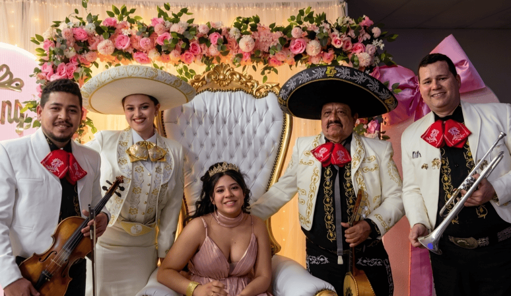 Cuatro músicos de Reyes del Mediterráneo con trajes de gala blancos y moños rojos posan junto a una joven quinceañera sentada en un trono decorado con flores.