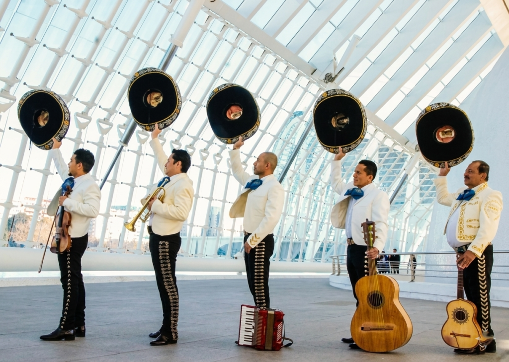 Cinco músicos mariachis de perfil alzando sus sombreros negros con la mano derecha en un gesto de saludo o celebración. Visten chaquetillas blancas con bordados dorados, moños azules y pantalones negros. Se encuentran en el interior de una estructura arquitectónica moderna de cristal y acero blanco.