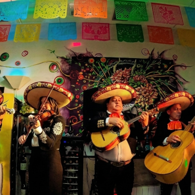 Cinco músicos del grupo Mariachi Reyes del Mediterráneo, incluyendo una mujer violinista, actúan en un local decorado con papel picado bajo luces festivas de colores.