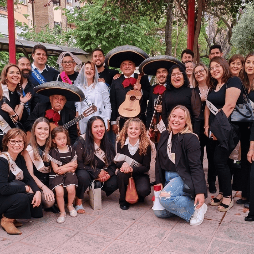 Integrantes de Reyes del Mediterráneo con trajes negros y sombreros posan en un parque rodeados por un grupo grande de mujeres que celebran una despedida de soltera con bandas de "Team Bride".
