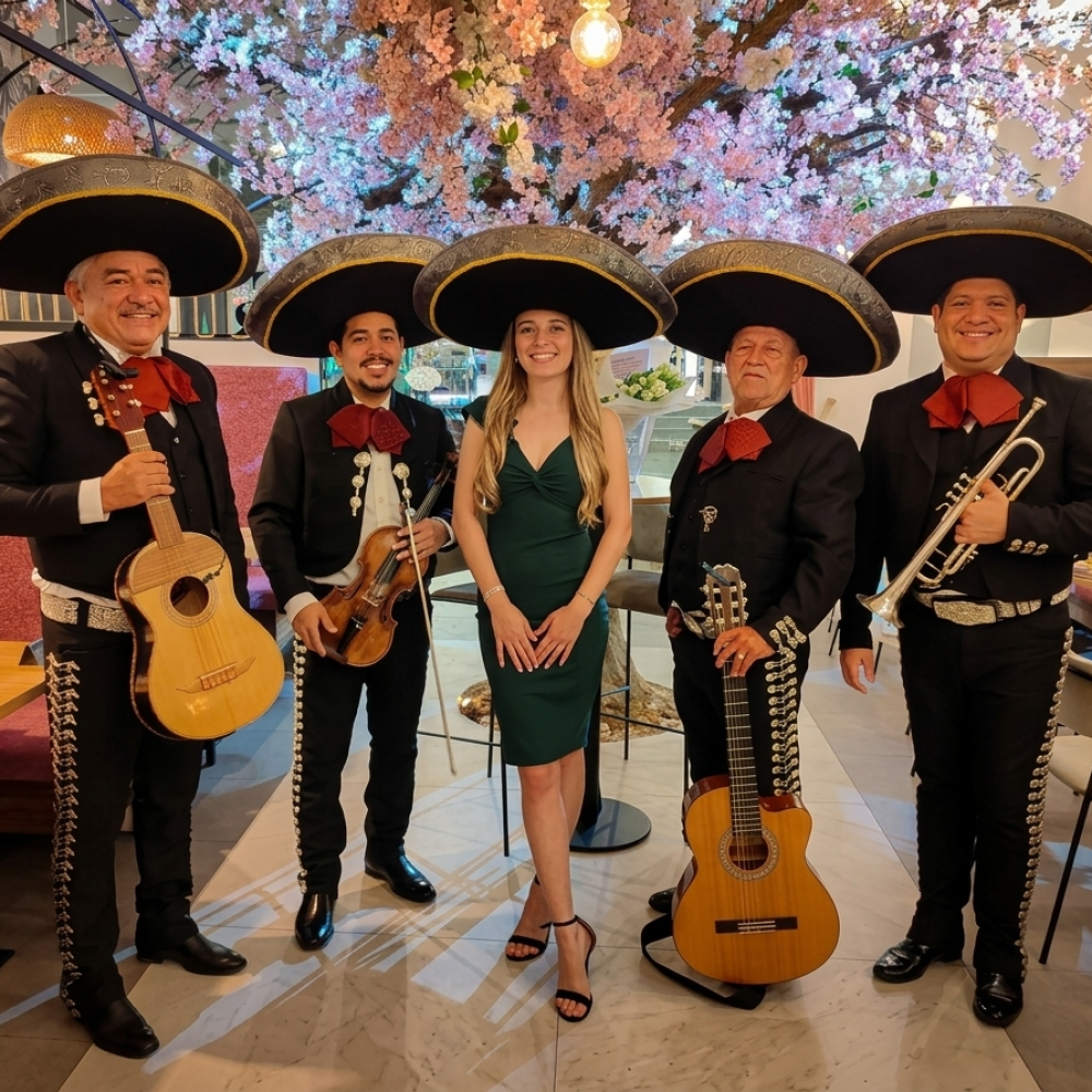 Cuatro integrantes del Mariachi Reyes del Mediterráneo con trajes negros y moños granates posando junto a una joven cumpleañera en un restaurante decorado con un árbol de flores rosas. Ella lleva un vestido verde oscuro y uno de los sombreros charros del grupo. Los músicos sostienen una vihuela, un violín, una guitarra y una trompeta.