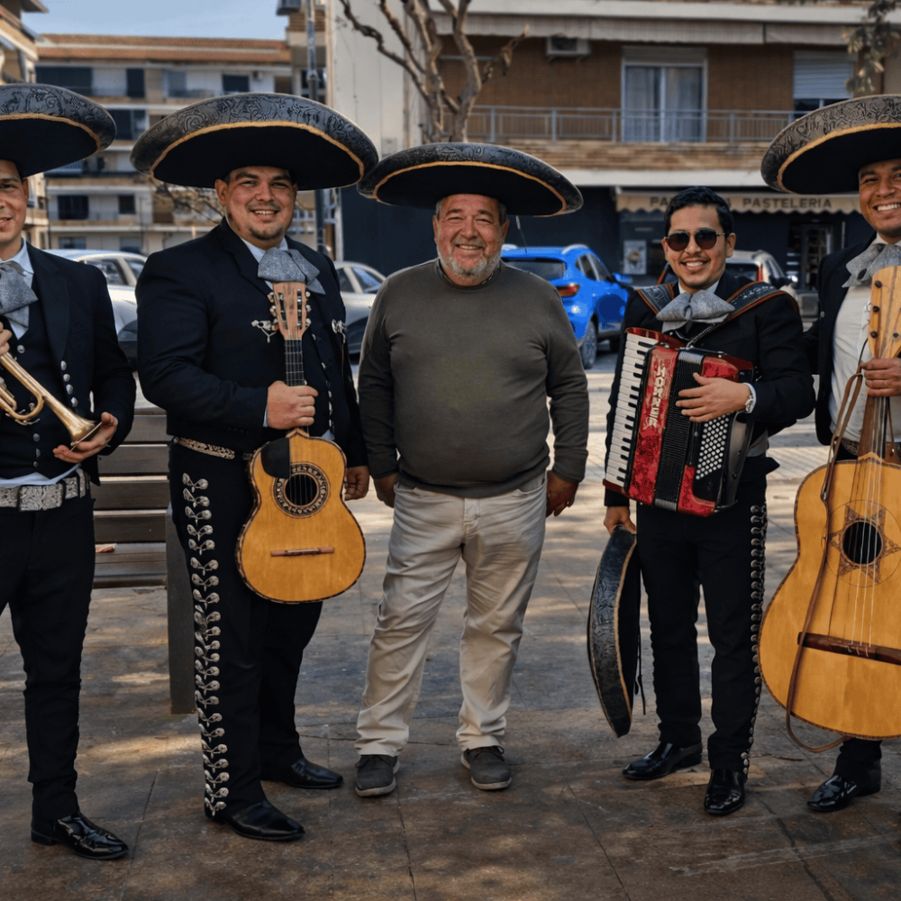 Tres músicos del Mariachi Reyes del Mediterráneo con trajes negros y moños azules posando junto a un hombre que celebra su cumpleaños. El cumpleañero sonríe mientras luce un sombrero charro negro y plata. Los músicos sostienen un violín, una trompeta y un guitarrón en un salón con decoración de globos dorados y azules de fondo.