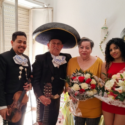 Cuatro mariachis con trajes de charro negros posan en el salón de una casa junto a una mujer mayor y una joven que sostienen ramos de flores durante una serenata sorpresa.