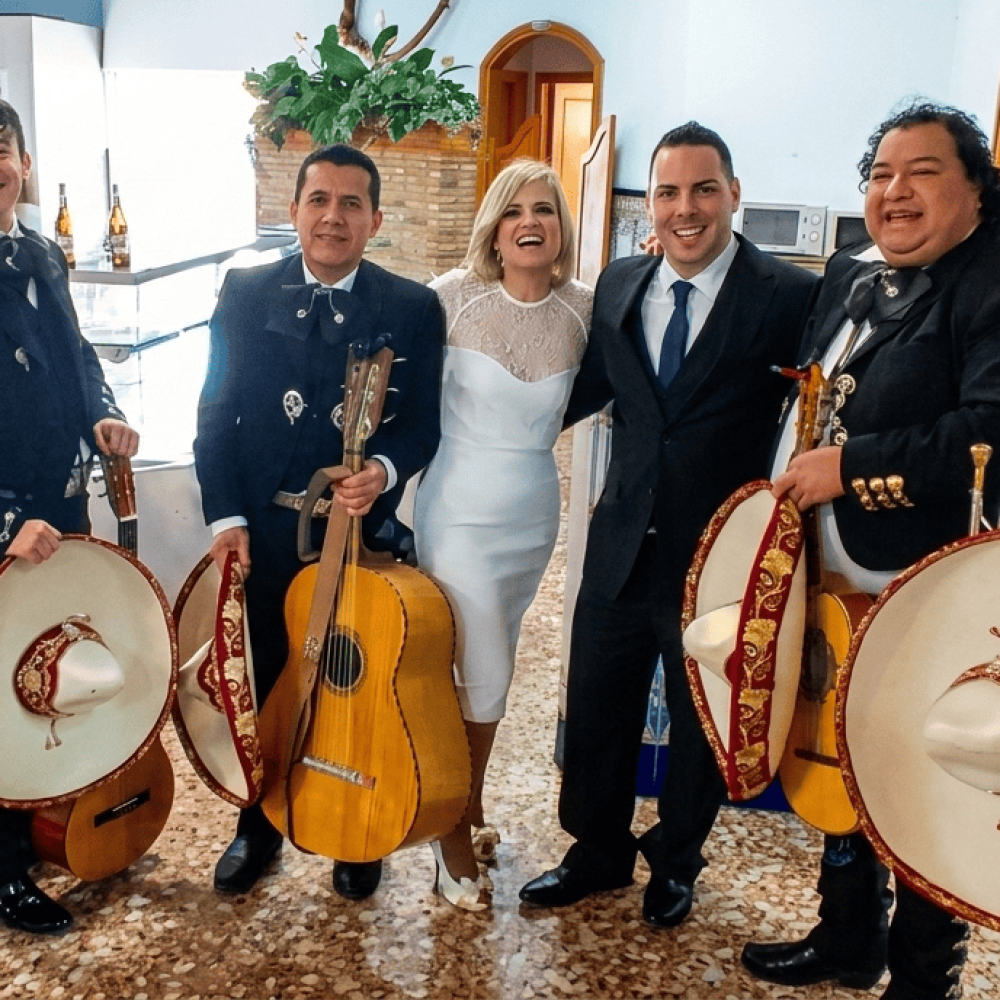 Cinco músicos del Mariachi Reyes del Mediterráneo con trajes negros y detalles dorados, posando junto a una pareja de novios en un salón. Los músicos sostienen sombreros charros blancos con bordados rojos y sus instrumentos tradicionales.