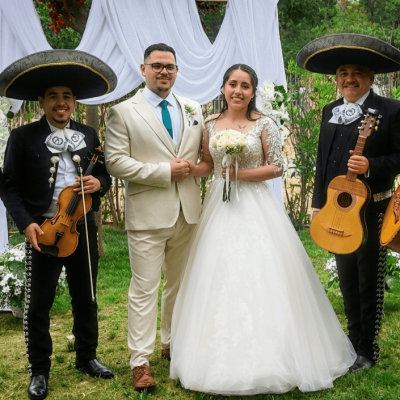 Cuatro músicos de Mariachi Reyes del Mediterráneo con traje de gala negro y moño blanco, posando junto a una pareja de novios bajo un arco floral blanco en un jardín. Los músicos sostienen una trompeta, un violín, una vihuela y un guitarrón. El novio viste traje crema con corbata verde y la novia un vestido blanco con encaje.
