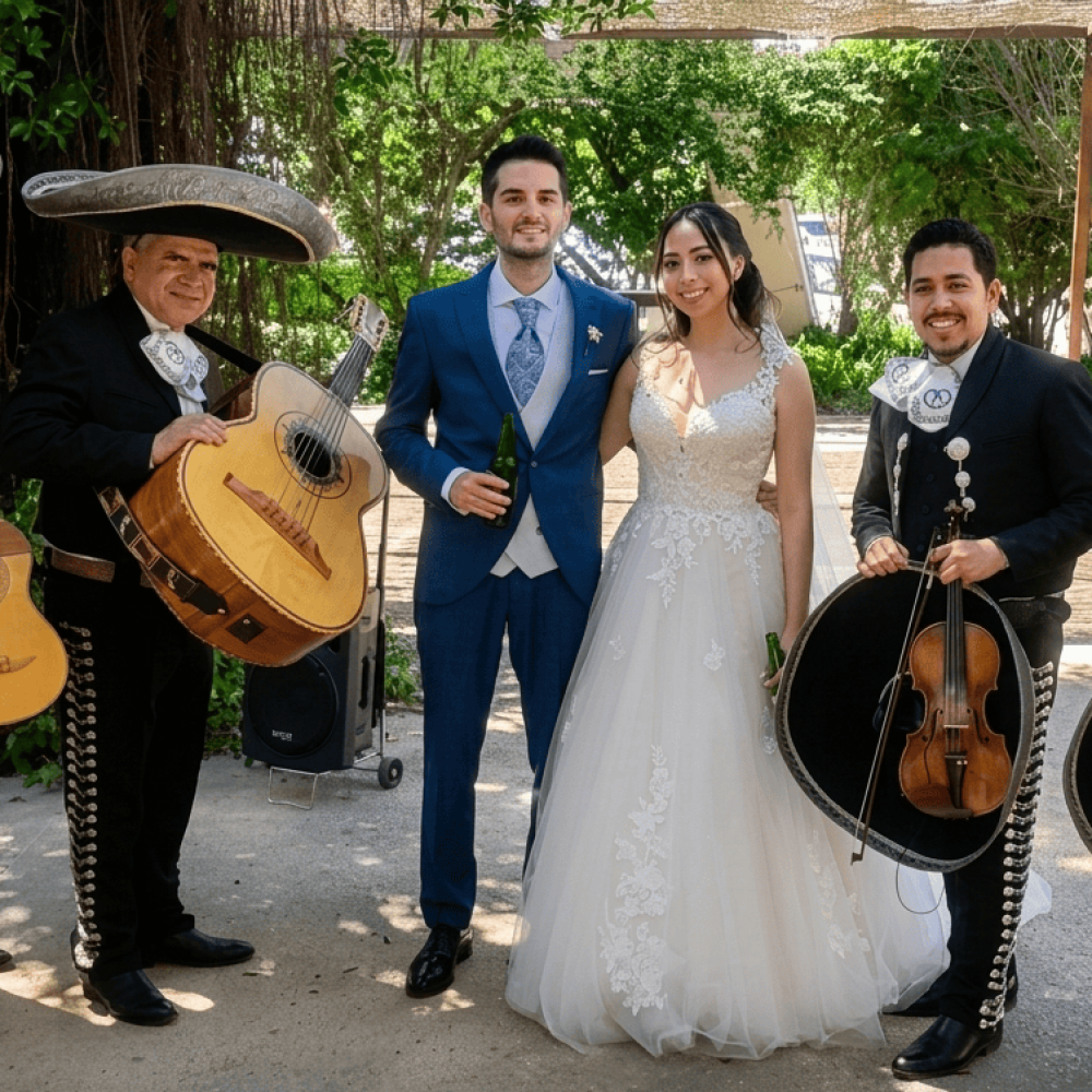 Cuatro integrantes del grupo Mariachi Reyes del Mediterráneo posando en un jardín exterior con una pareja de novios recién casados. Los músicos visten trajes de gala negros con moños blancos bordados. Sostienen una vihuela, un guitarrón, un violín y una trompeta. El novio viste traje azul y la novia un vestido blanco con encaje.