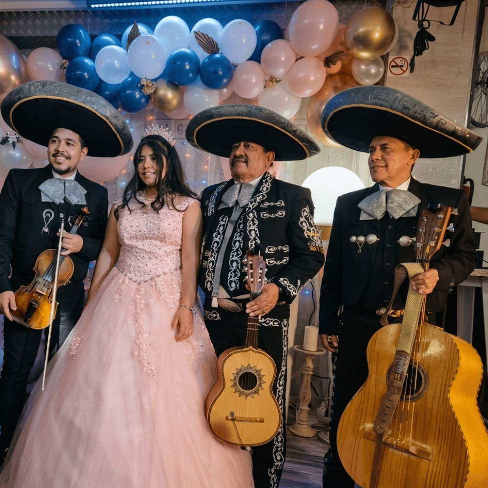 Tres músicos del Mariachi Reyes del Mediterráneo con trajes de gala negros y plata, posando junto a una joven quinceañera con un vestido rosa de princesa y tiara. De fondo, una decoración de globos azules, blancos y dorados. Los músicos sostienen un violín, una vihuela y un guitarrón.