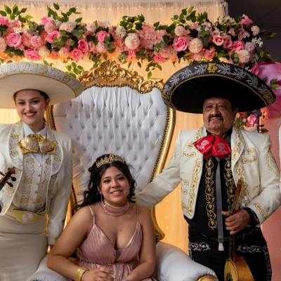 Cuatro músicos de Reyes del Mediterráneo con trajes de gala blancos y moños rojos posan junto a una joven quinceañera sentada en un trono decorado con flores.