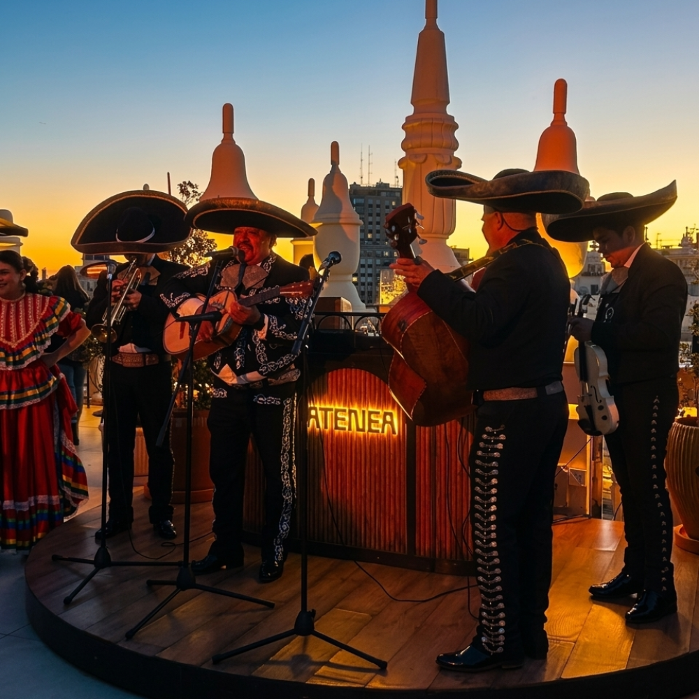 Grupo de mariachis actuando en una terraza al aire libre durante el atardecer, con el cielo naranja de fondo y estructuras arquitectónicas blancas.