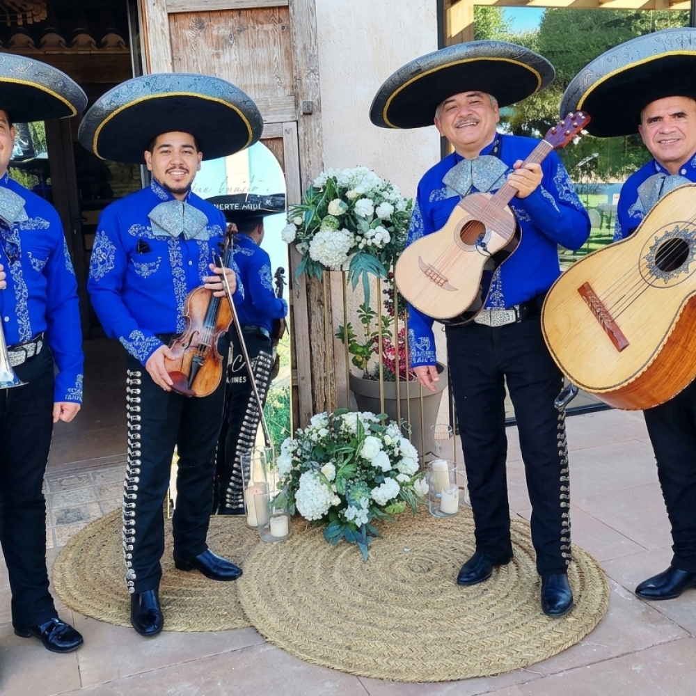 Cuatro músicos de Mariachi Reyes del Mediterráneo posando en una boda. Llevan camisas azul eléctrico con bordados plateados, moños grises, sombreros charros negros y pantalones con botonadura lateral. Sostienen una trompeta, un violín, una vihuela y un guitarrón frente a un decorado floral blanco.