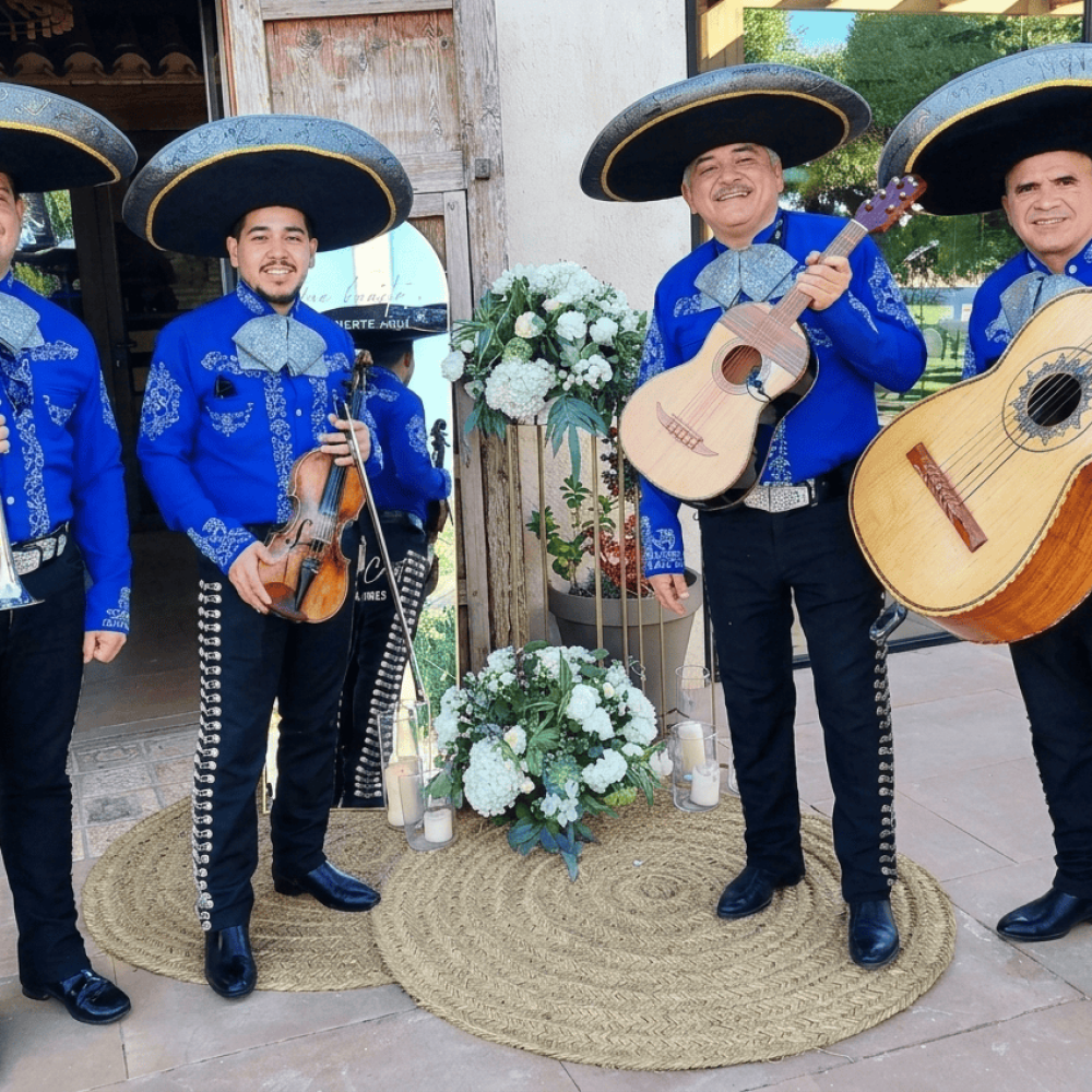 Mariachis en Elche | Serenatas, Bodas y Eventos 24H 53 Cuatro músicos de Mariachi Reyes del Mediterráneo posando en una boda. Llevan camisas azul eléctrico con bordados plateados, moños grises, sombreros charros negros y pantalones con botonadura lateral. Sostienen una trompeta, un violín, una vihuela y un guitarrón frente a un decorado floral blanco.