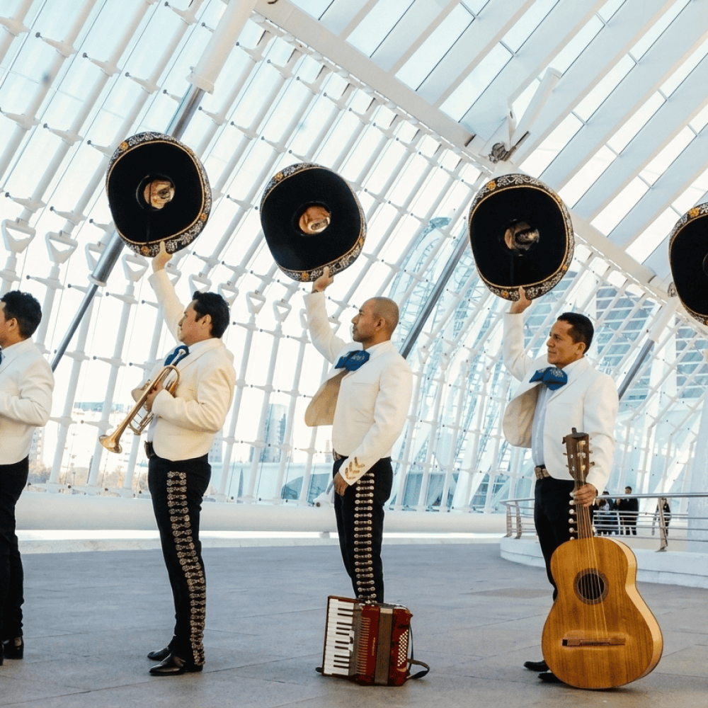 Mariachis en Elche | Serenatas, Bodas y Eventos 24H 55 Cinco músicos mariachis de perfil alzando sus sombreros negros con la mano derecha en un gesto de saludo o celebración. Visten chaquetillas blancas con bordados dorados, moños azules y pantalones negros. Se encuentran en el interior de una estructura arquitectónica moderna de cristal y acero blanco.