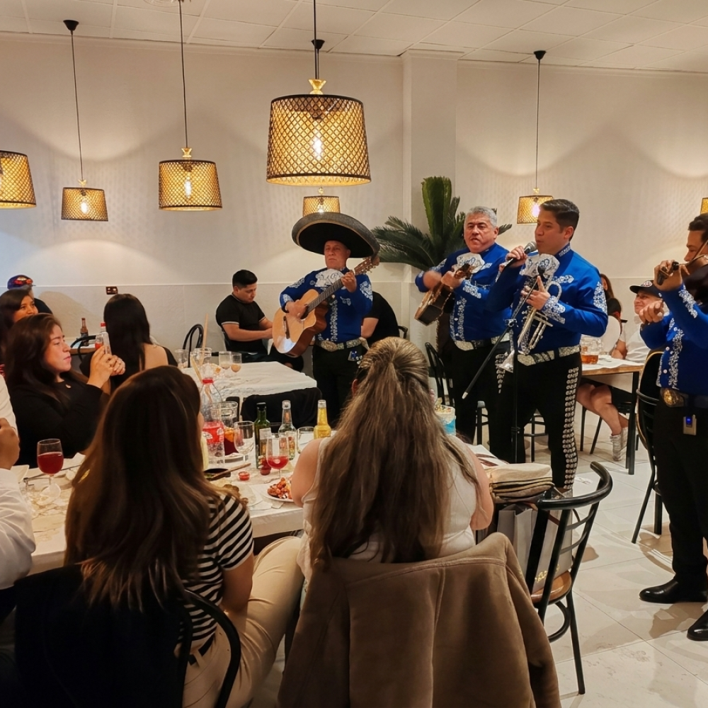Cuatro integrantes del Mariachi Reyes del Mediterráneo con trajes azul eléctrico actuando en directo en un restaurante. Los músicos tocan la guitarra, trompeta y violín mientras los comensales disfrutan de la cena bajo lámparas de mimbre modernas. El ambiente es acogedor y animado.
